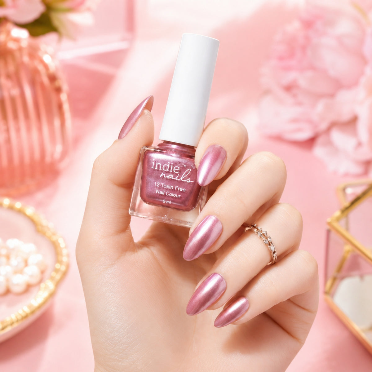 A hand with metallic pink nails holds a bottle of Indie Nails nail polish in a matching shimmering pink shade, set against a soft pink floral background.
