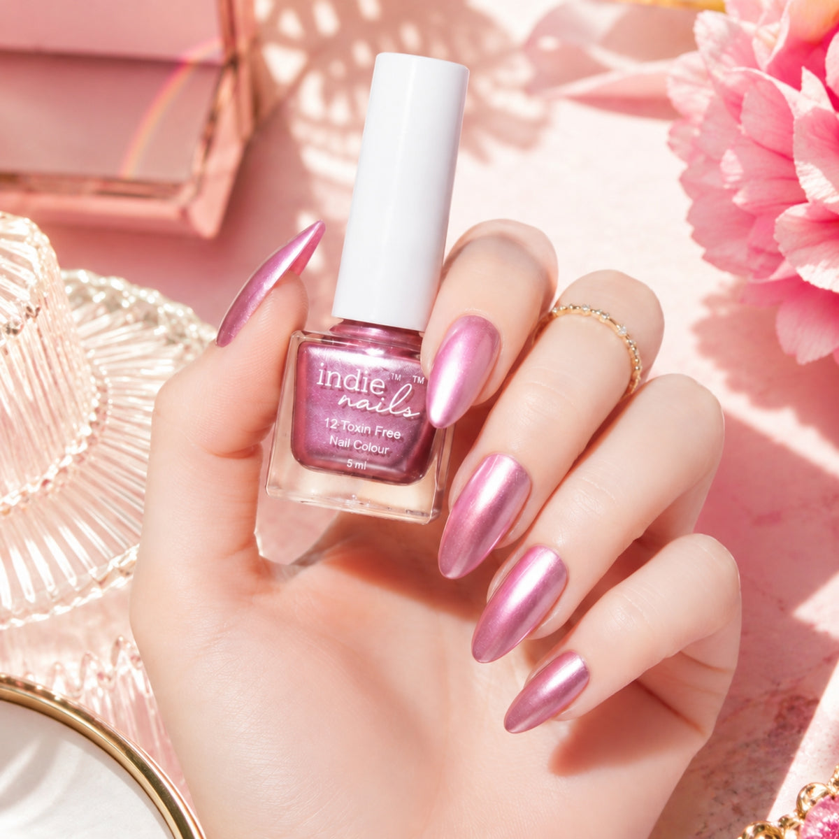 A hand with metallic pink manicured nails holds a bottle of Indie Nails 12 Foam Free Gel Nail Color in a shimmering pink shade, styled with pink flowers and a pink hat on a pink background.