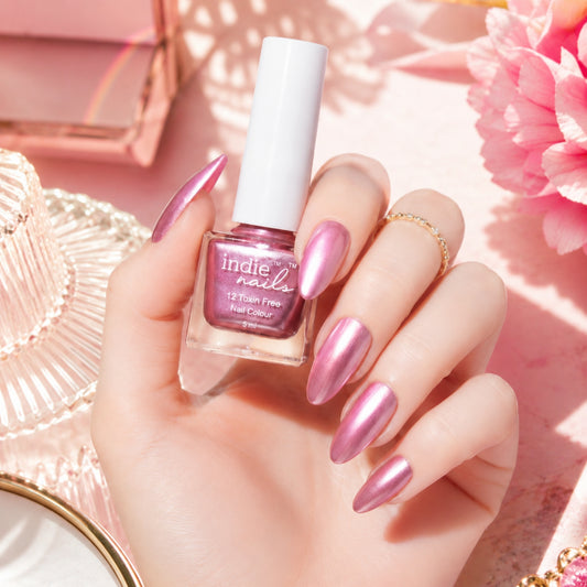A hand with metallic pink manicured nails holds a bottle of Indie Nails 12 Foam Free Gel Nail Color in a shimmering pink shade, styled with pink flowers and a pink hat on a pink background.