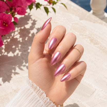A hand with long, metallic pink stiletto-shaped nails, wearing a white textured sweater and a gold ring, posed outdoors against a sunlit stone wall with pink flowers in the background.