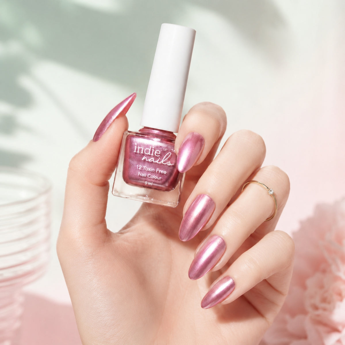 A hand with long, metallic pink nails holds a bottle of Indie Nails metallic pink nail polish against a soft, light pink and white background with subtle leaf shadows.