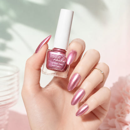 A hand with long, metallic pink nails holds a bottle of Indie Nails metallic pink nail polish against a soft, light pink and white background with subtle leaf shadows.