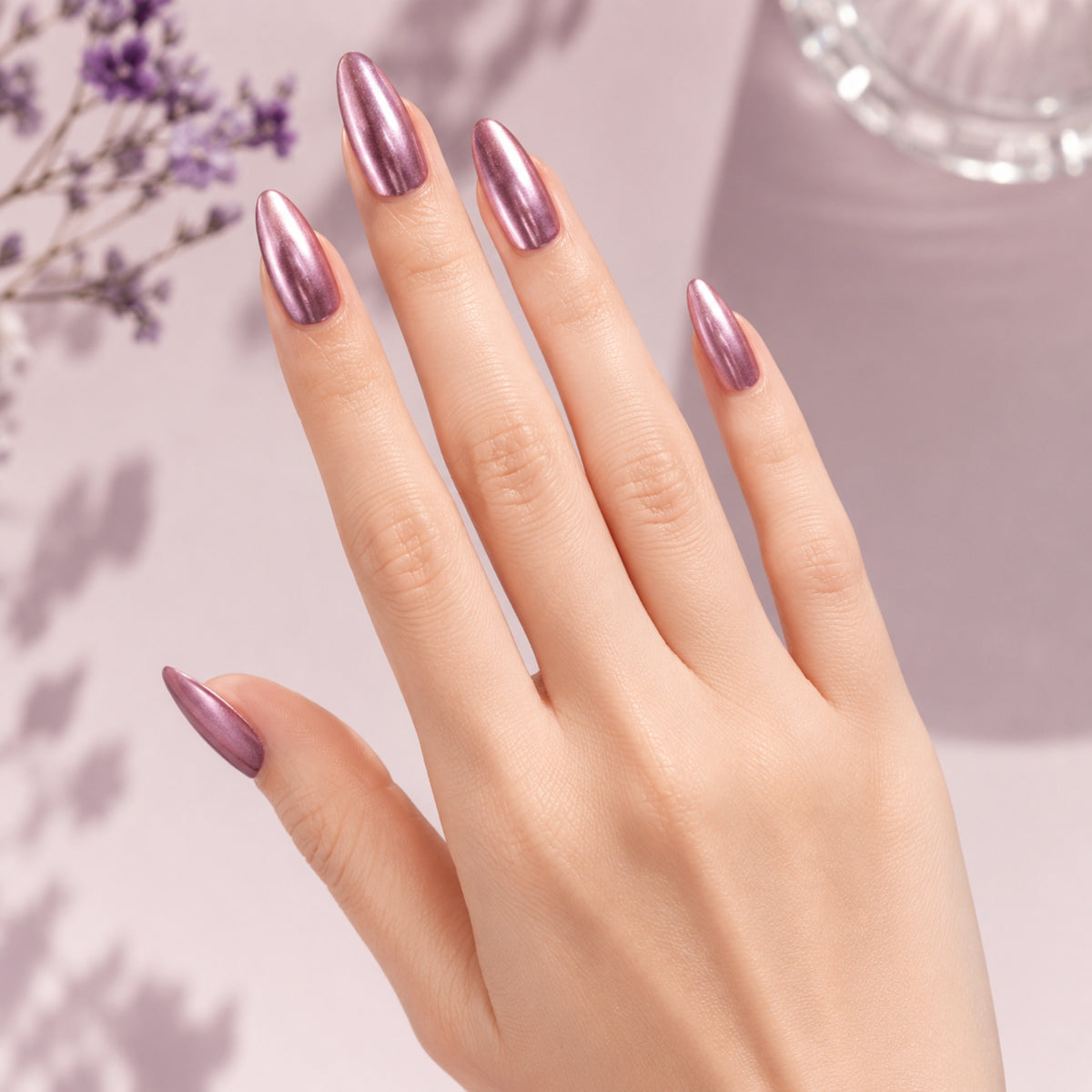 A close-up of a hand with long, almond-shaped nails painted in a metallic, shimmery mauve-purple nail polish, set against a soft lavender background with decorative dried flowers.